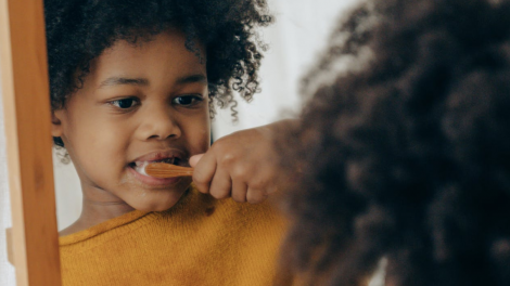Un bambino di circa quattro anni si guarda allo specchio mentre si lava i denti da solo con uno spazzolino manuale. Indossa una maglietta gialla, dello stesso colore dello spazzolino.
