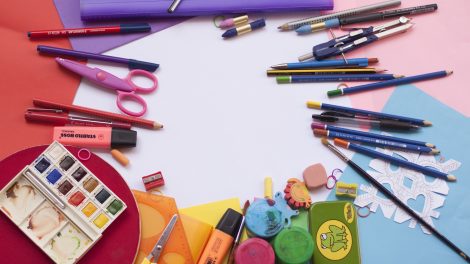 Su una scrivania sono posati dei materiali di cancelleria colorati, in modo che lascino uno spazio vuoto centrale. Sono presenti forbici dalla punta arrotondata, pastelli, matite, gomma da cancellare, colori e righelli