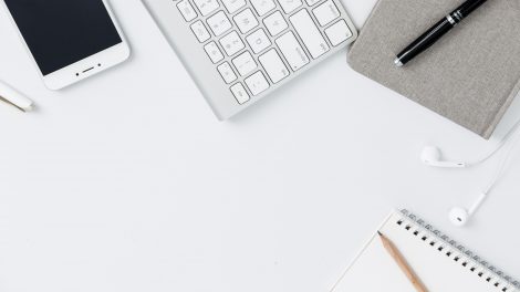 Una scrivania vista dall'alto con sopra riposti uno smartphone, una tastiera per computer, un'agenda con una penna, degli auricolari e un quadernetto