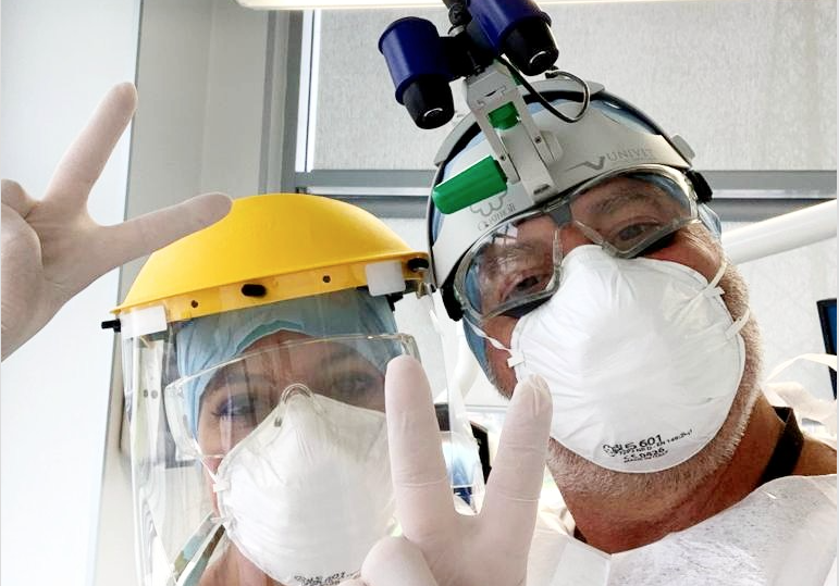 Un dentista e la sua assistente salutano in camera facendo il gesto della vittoria, completamente bardati con i dispositivi di protezione individuali richiesti dalle misure di prevenzione contro la diffusione del covid19