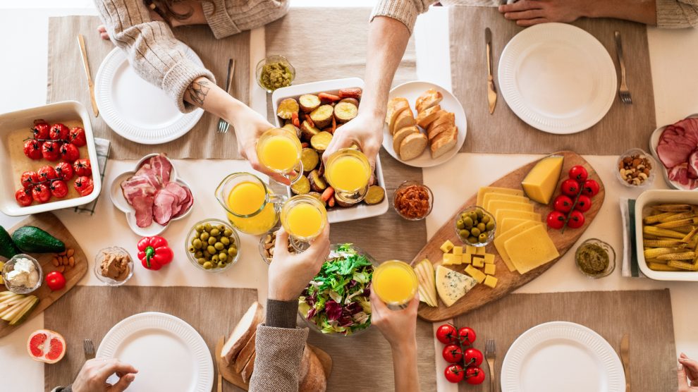 una tavolata imbandita di cibo (pomodori, olive, formaggi, pane, grissini e insalata) inquadrata dall'alto. Al tavolo quattro persone che stanno facendo un brindisi con un succo di frutta