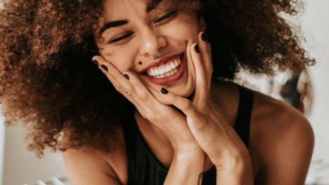 Una ragazza riccia con piercing al naso e al labbro sorride, tenendo il viso tra le mani