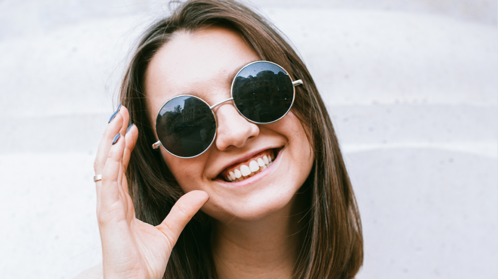 Una ragazza sorride mentre indossa un paio di occhiali da sole scuri con lenti tonde e molto grandi